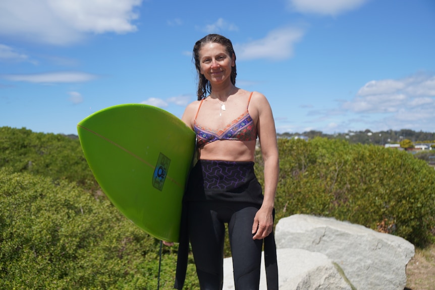 A woman smiles at a camera half in a wetsuit and bikini with a green surfboard.