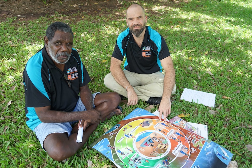 An Indigenous man and non-Indigenous man sitting in the grass with a colourful board.