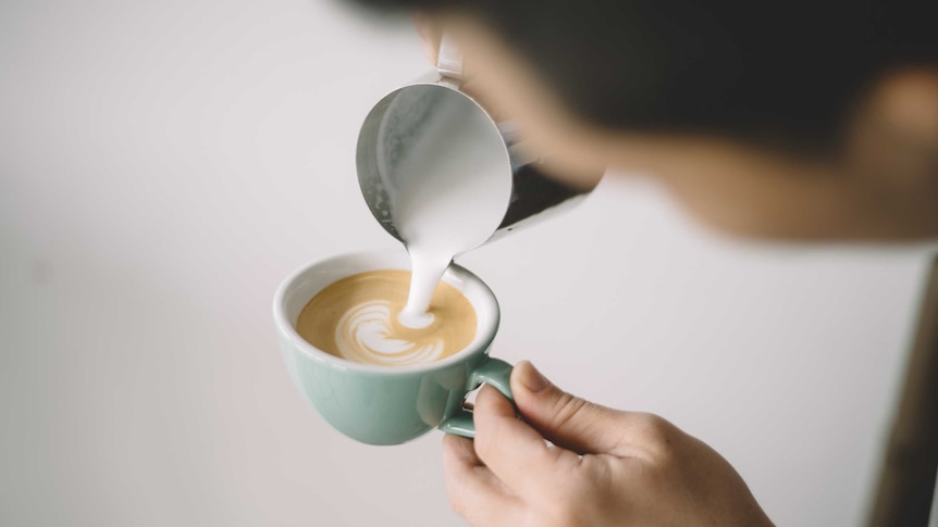 A man pours milk into a white ceramic mug containing coffee.