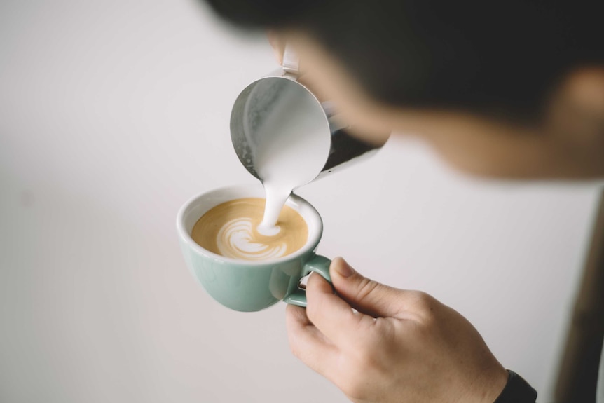 A man pours milk into a white ceramic mug containing coffee.