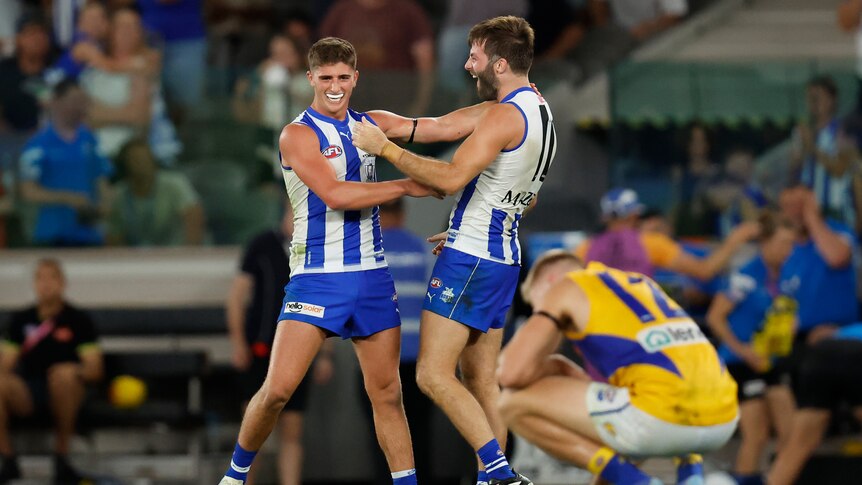 Two North Melbourne players hold each other and shout in triumph after a game as an opposing player crouches on the ground.