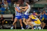 Two North Melbourne players hold each other and shout in triumph after a game as an opposing player crouches on the ground.
