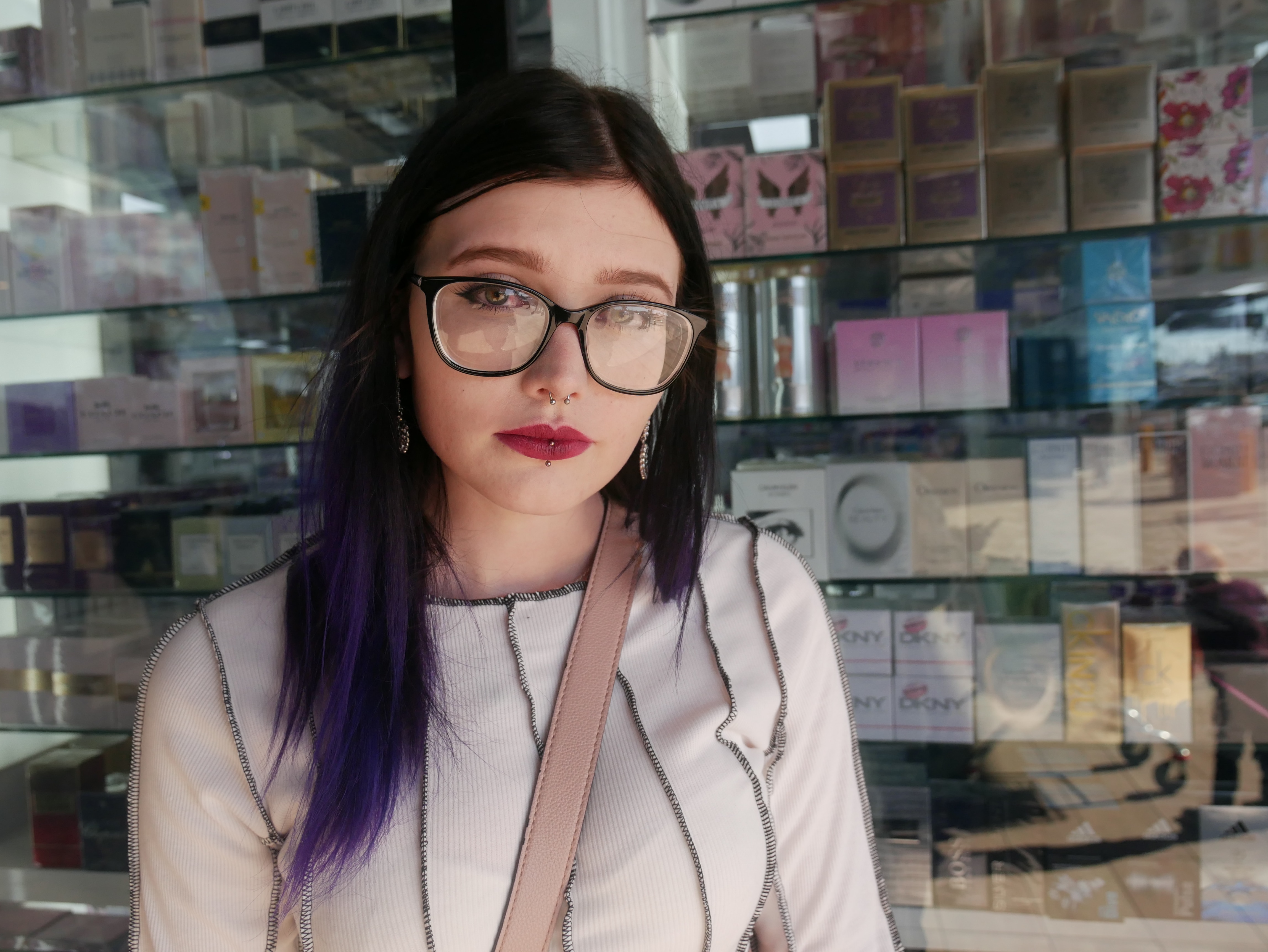 A woman with glasses and long dark hair stands in front of a shop window.