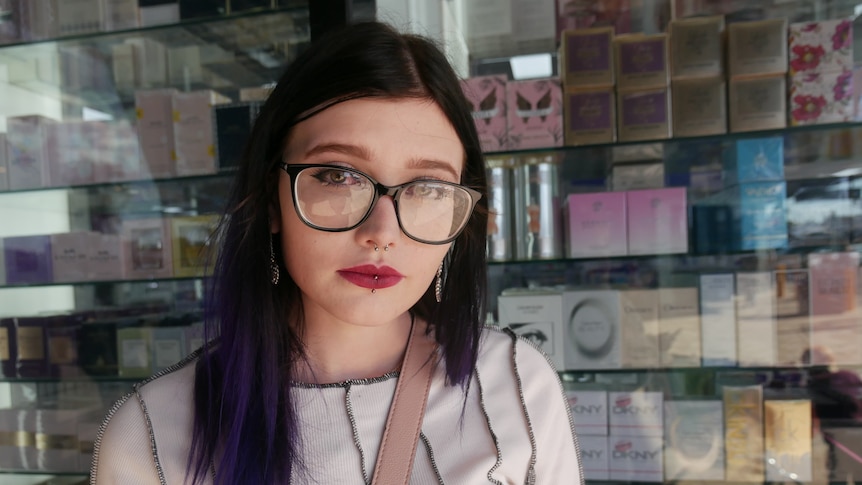 A woman with glasses and long dark hair stands in front of a shop window.