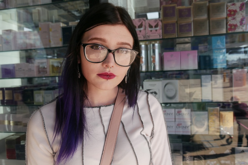 A woman with glasses and long dark hair stands in front of a shop window.