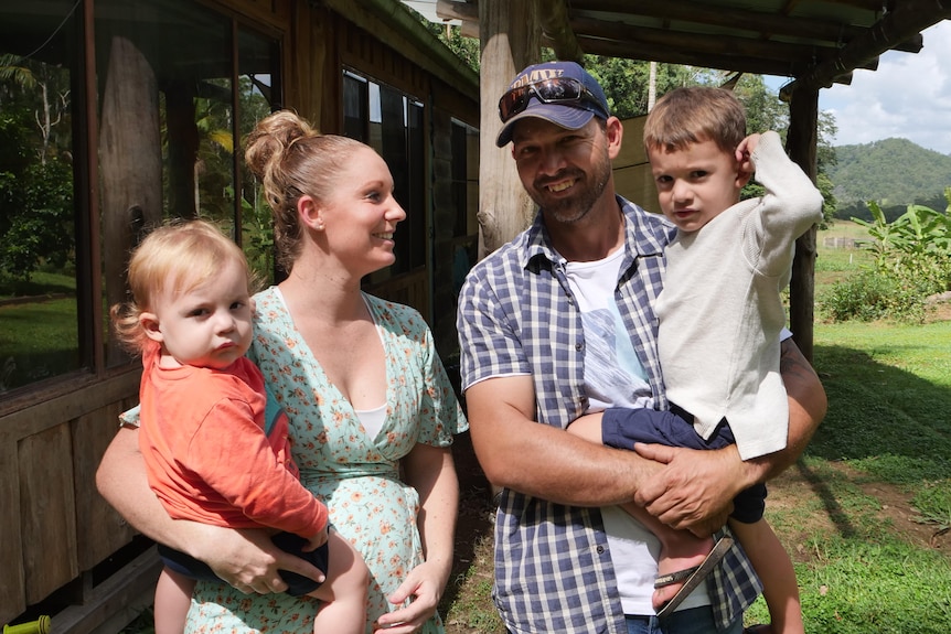 A man and a pregnant woman stand together outside a cabin, each holding a young child.