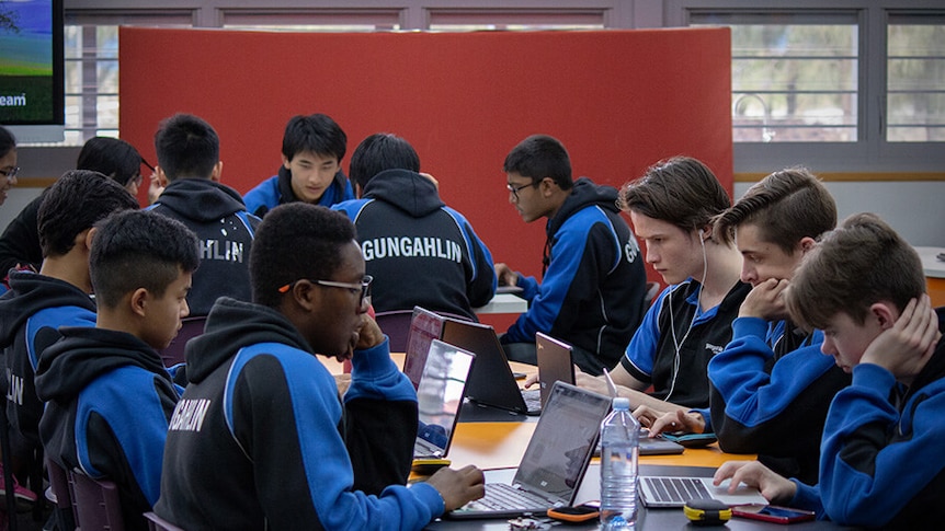 Boys in black and blue uniform sit at desks using laptop computers.
