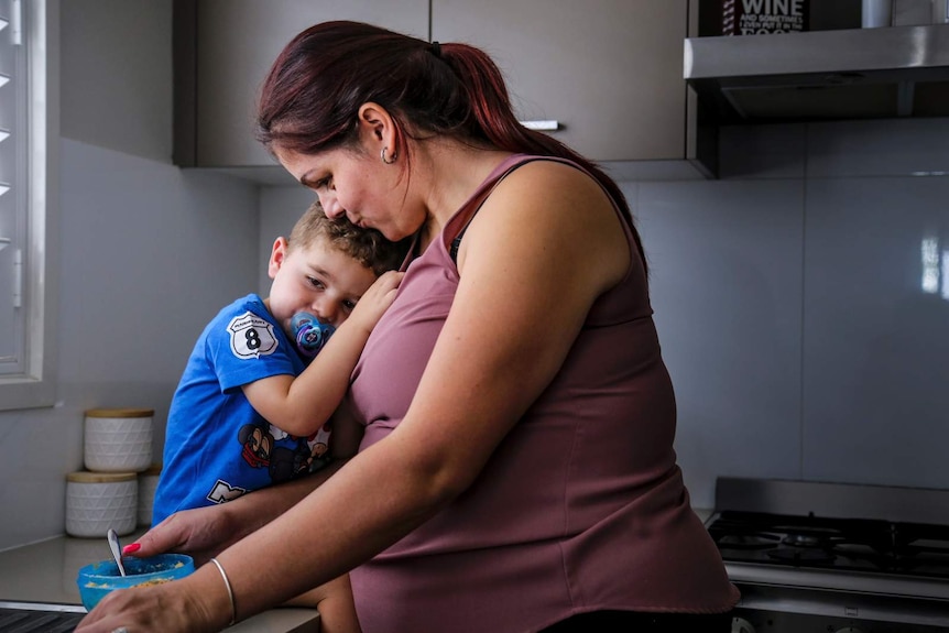 a young boy sitting on a kitchen bench cuddling his mother who is making him cereal