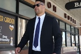 A man in his 30s with short hair wearing dark glasses and a dark suit pulls a suitcase along a city street.