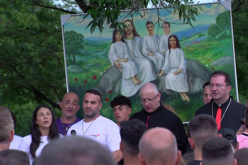 A crowd, including Leila and Danny Abdallah, gathers to pray in front of a painting of four dead children sitting with Jesus.