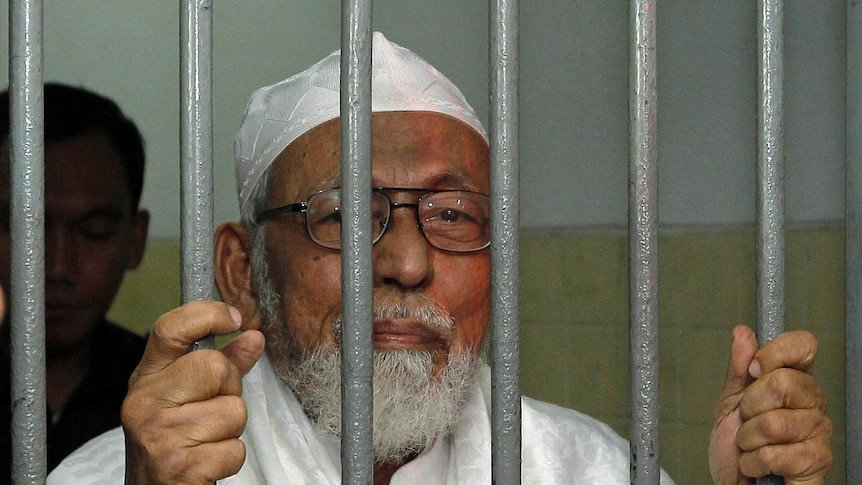 An elderly man stands behind bars while holding them, and wearing glasses.