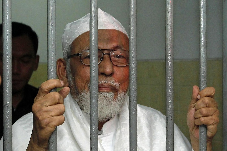 An elderly man stands behind bars while holding them, and wearing glasses.