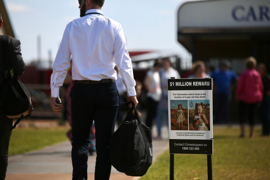  A man in a shirt and pants walks past a poster at carnarvon airport