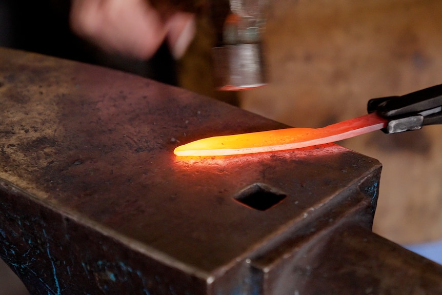 A flaming hot knife is in the forge, being hit with a hammer.