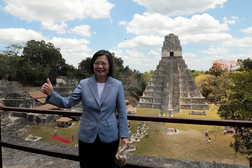 Tsai Ing-wen, wearing a blue linen blazer, stands in front of a rail above Mayan ruins, giving a thumbs up