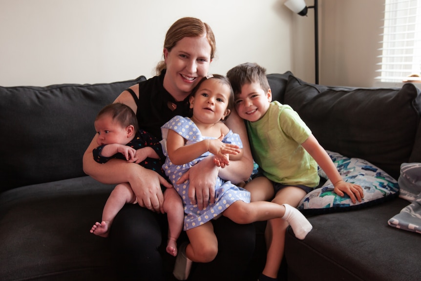 A mom with two toddlers and a newborn smile while sitting on the couch 