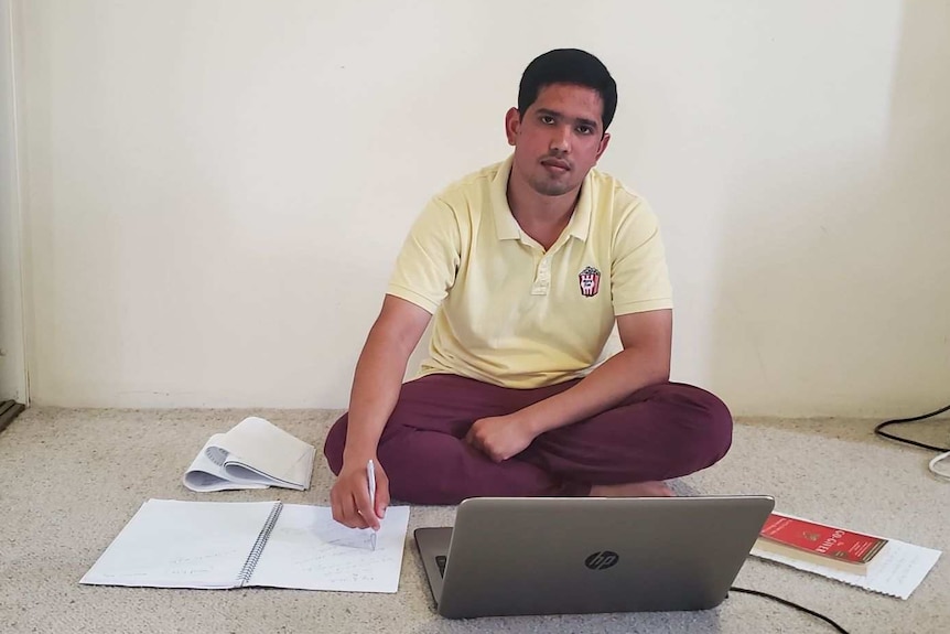A young man sits cross-legged on a bedroom floor with his laptop and notebooks open in front of him