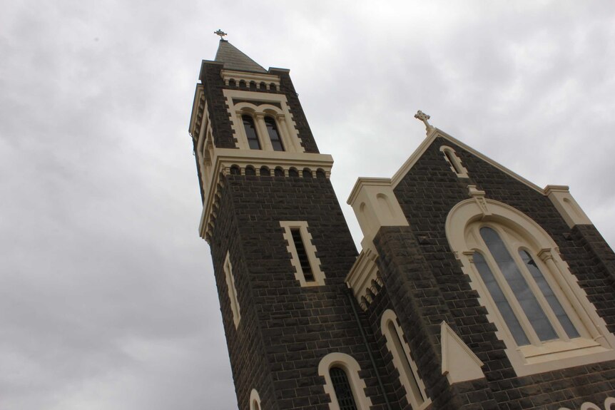 The facade of a church with dark brown bricks and light brown detail. A cloudy sky is in the background.