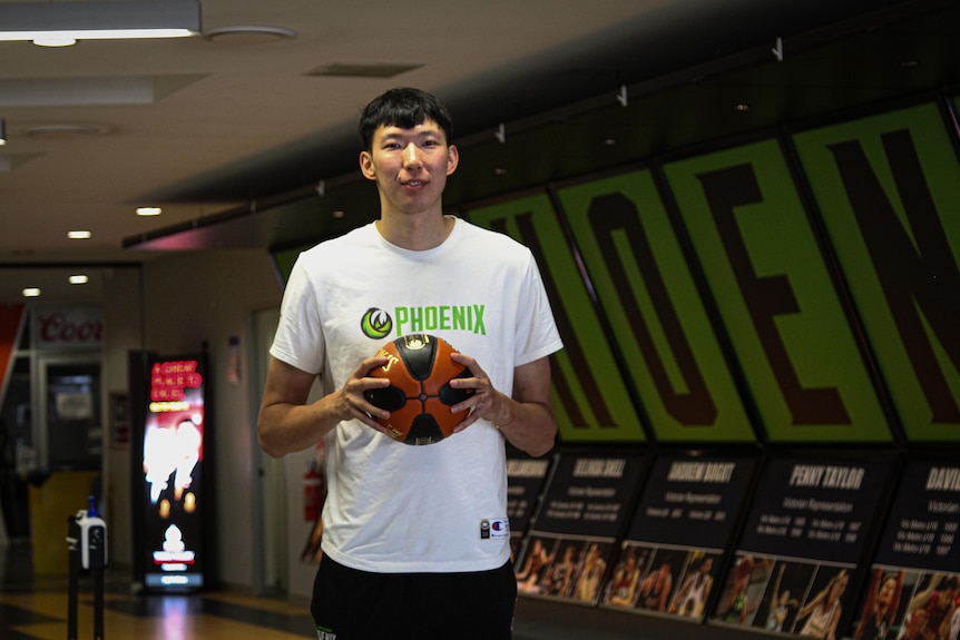 A tall man wearing a team-branded shirt stands holding a basketball.