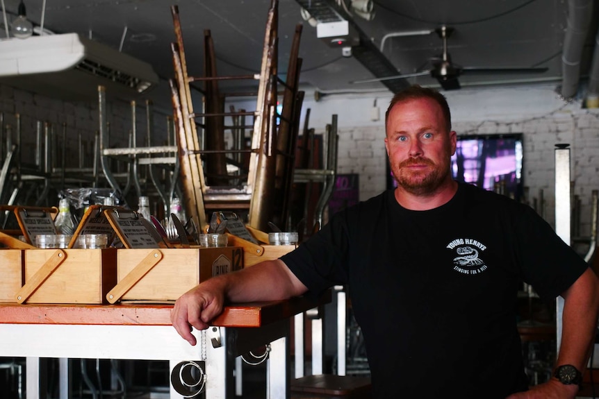 A serious-looking man in a black t-shirt with a small white logo stands in a room with chairs, boxes, stacked on tables.