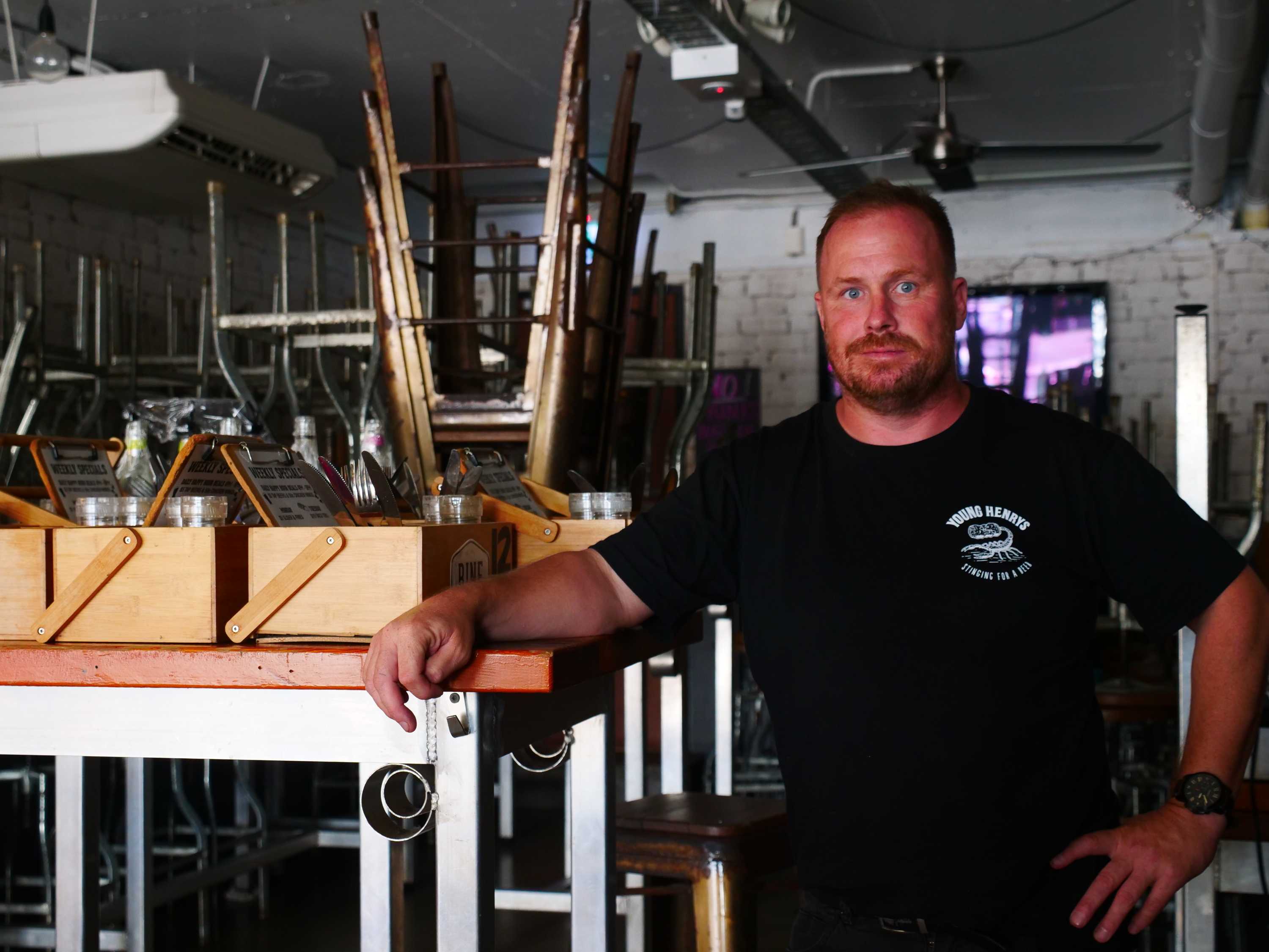 A serious-looking man in a black t-shirt with a small white logo stands in a room with chairs, boxes, stacked on tables.