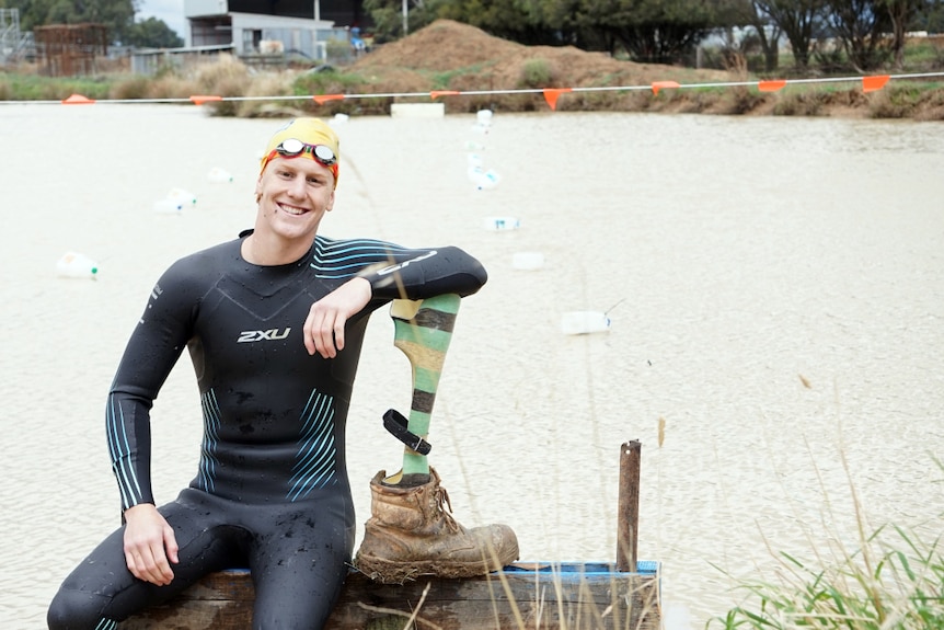 A smiling young man in a wetsuit and swimming cap sits on the banks of a large dam with makeshift lane ropes.
