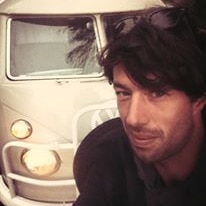 A man squats in front of a Volkswagen Kombi car behind him as he smiles at the camera.