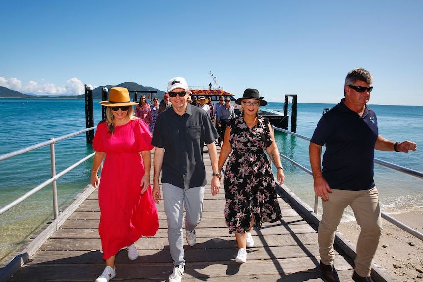 Four men and woman people walk off a boat towards land in the backdrop of turquoise waters.