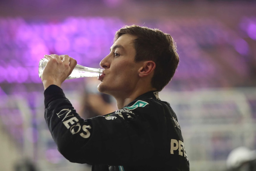 George Russell takes a swig out of a bottle of water. Purple lights fill the background