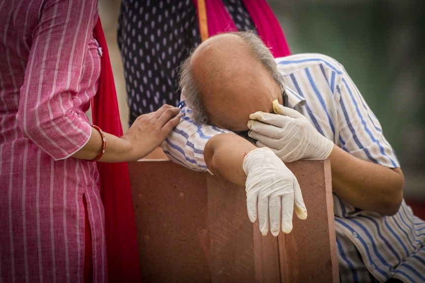 A man in gloves leans on a chair and covers his face as a woman rests a hand on his shoulder