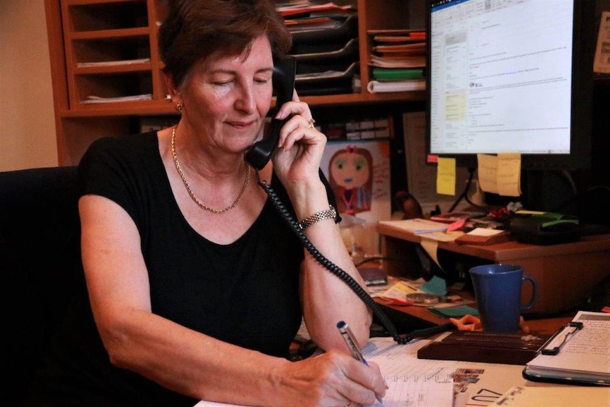 A middle-aged woman sits holding a pen and talking into a landline phone receiver in an office next to a desk and computer.