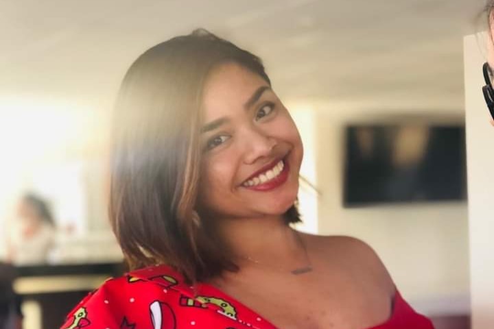 A smiling, dark-haired Asian woman in a red, patterned dress with bright sunshine streaming through the window behind her