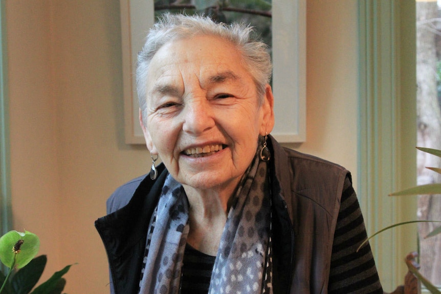An elderly woman with short grey hair smiles at the camera inside her living room.