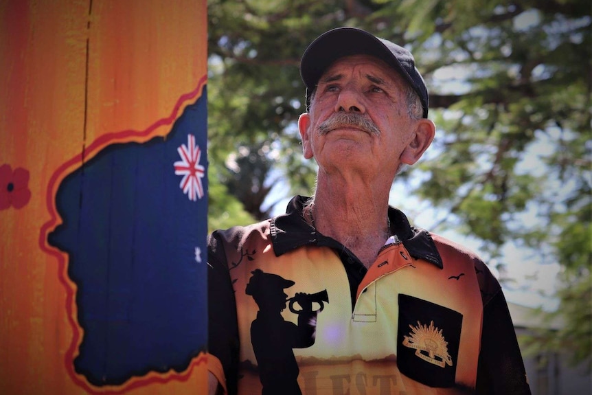 Man with moustache and wearing an Anzac day shirt stands next to a painted power pole and gazes into the distance