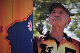 Man with moustache and wearing an Anzac day shirt stands next to a painted power pole and gazes into the distance