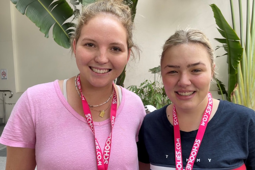 Two girls with blonde hair standing next to each other smiling, both wearing pink Schoolies lanyards
