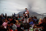A masked woman holding a baby stands on crowded boat with smoldering volcano behind