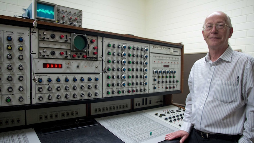 A man stands next to a large grey cabinet covered in dials and switches.