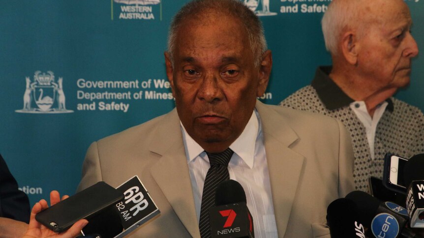 An elderly man in a suit speaks to media at a press conference at Consumer Protection offices.