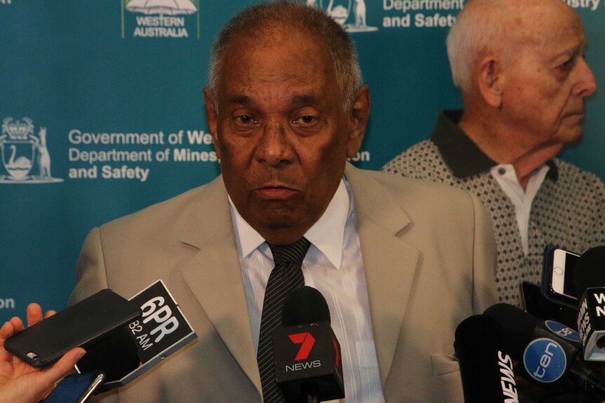 An elderly man in a suit speaks to media at a press conference at Consumer Protection offices.