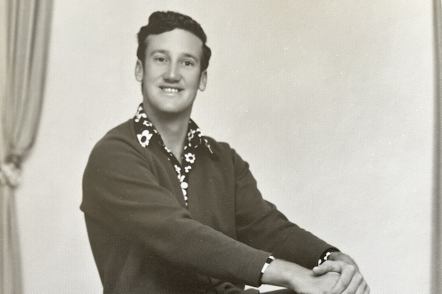 A young brunette man sits on a chair wearing a floral shirt and cardigan, with his hands on his knees, smiling at the camera.
