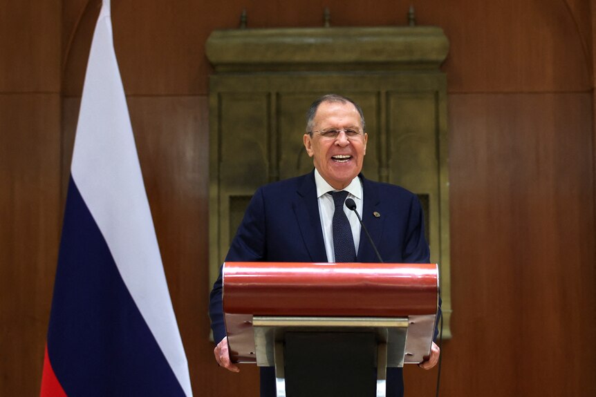 An older man in a blue suit laughs behind a lectern in front of a Russian flag.