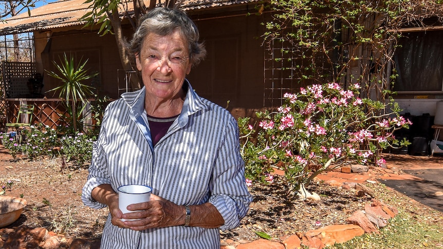 An older woman stands in the shade of a tree out the front of a bungalow.