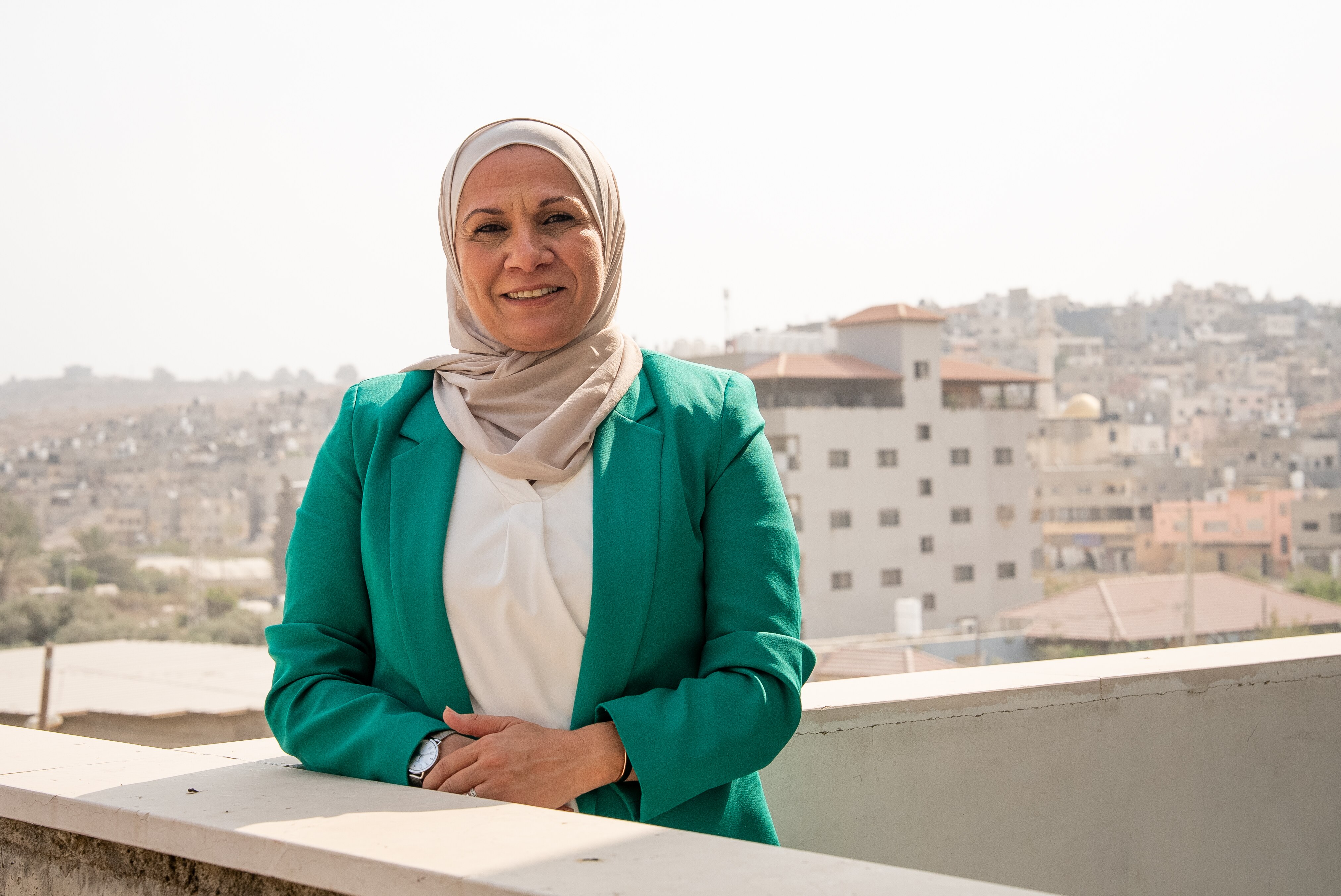 A woman wearing a hijab and a green jacket leans against a railing on a rooftop.