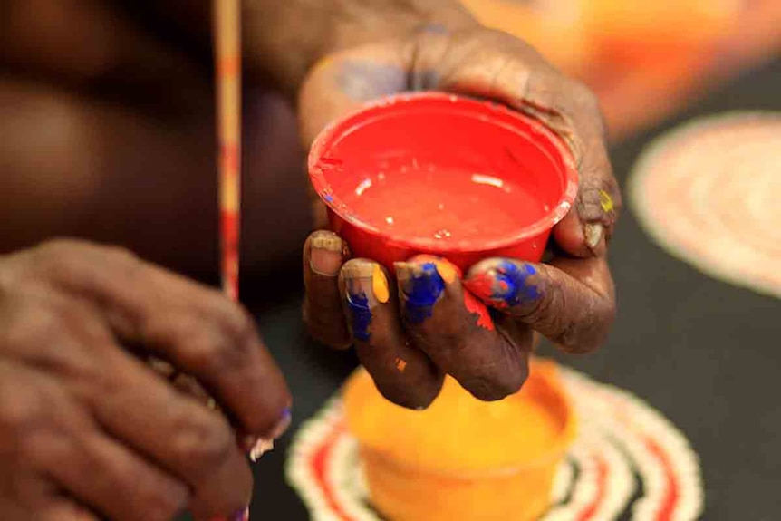 Indigenous woman dips her paint brush into a bucket with red paint.
