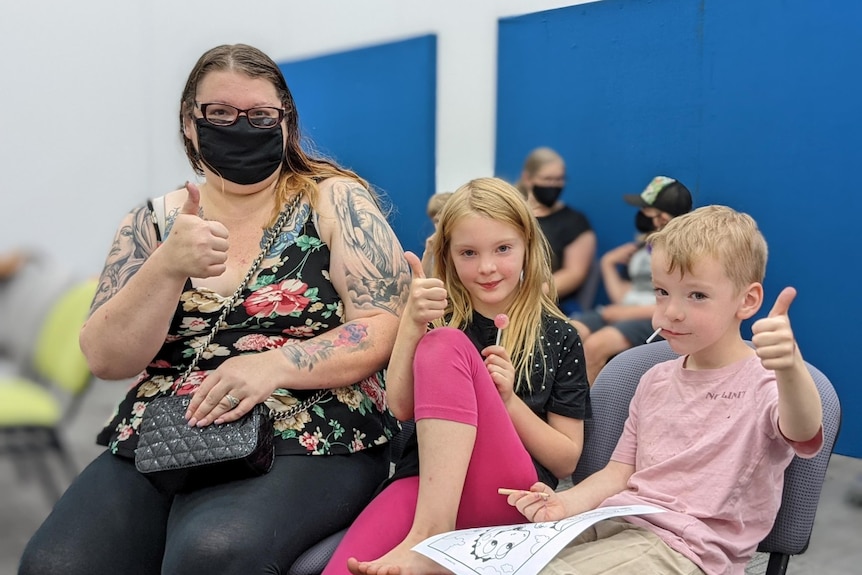 A woman wearing a mask gives the thumbs up with her two children 