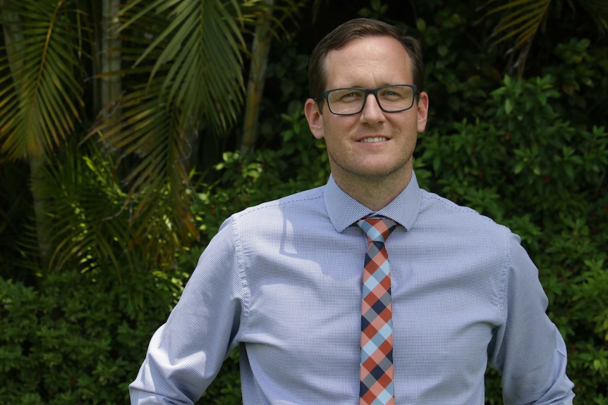 A dark-haired, bespectacled man in a shirt and tie, smiling with his hands on his hips.