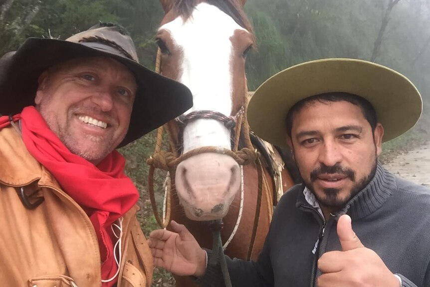 Two men stand either side of a brown horse, smiling and giving a thumbs up to the camera