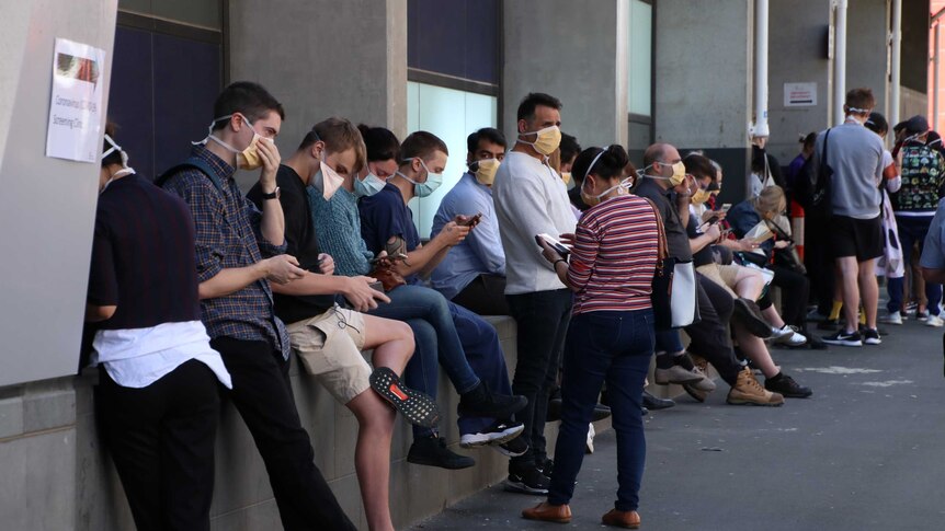 About a dozen people wearing masks sitting outside looking at their phones in a queue.