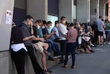 About a dozen people wearing masks sitting outside looking at their phones in a queue.